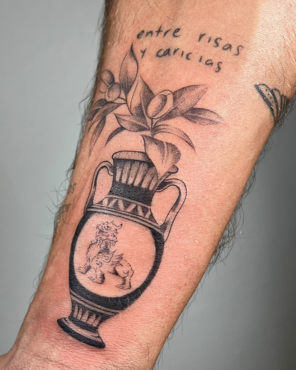 Jarrón griego con flores y la frase «entre risas y caricias» en el antebrazo